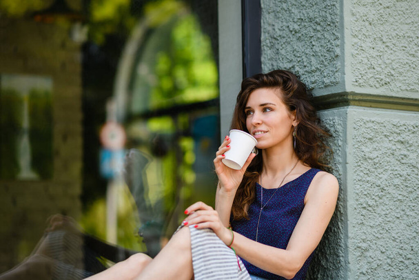 Young attractive woman enjoying takeaway coffee while sitting by a huge window outdoors. The girl is holding a disposable cup. - Photo, image