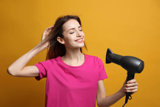 Beautiful young woman using hair dryer on yellow background - 写真・画像