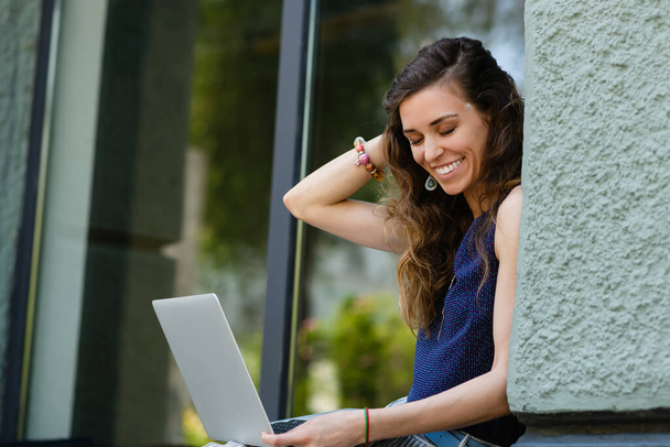 Young attractive woman with laptop sits on the windowsill outdoors. The girl straightens her hair with her hand. - Foto, afbeelding