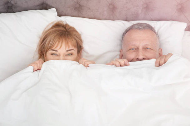 Mature couple hiding together under blanket in bed at home - Fotó, kép