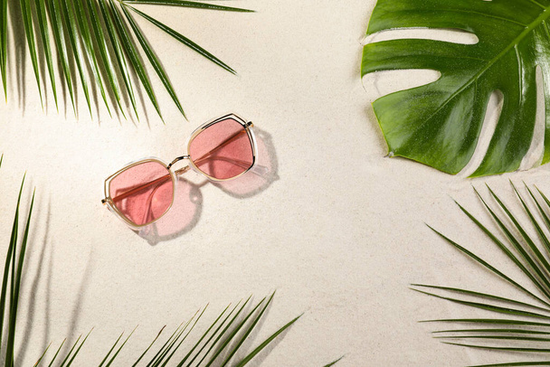 Stylish sunglasses and different tropical leaves on sand, flat lay - Фото, зображення