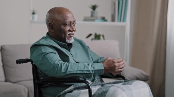 Old 60s African senior mature man alone older grandfather in wheelchair indoor at home looking window thinking pondering pensive contemplate look at camera smiling posing at apartment health insurance - Filmati, video