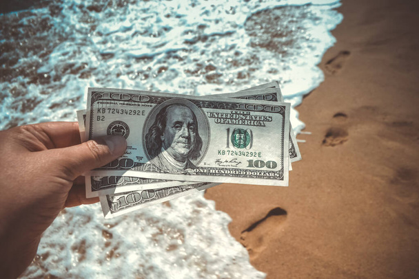 Girl holding money bill of 300 dollars on background of sea ocean waves with white foam and sand wet beach with footprints in sand close-up. Concept finance money holiday traveling dollars vacation - Foto, Bild
