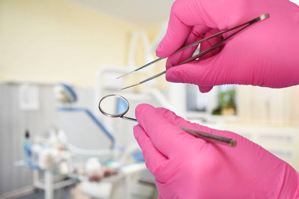 Dentist's hands in pink latex gloves with a tweezers and a mouth mirror with a dental clinic on the background. Medical tools concept. - Fotó, kép