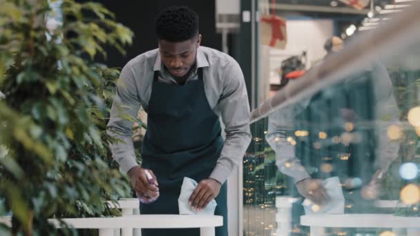 Young man waiter in apron does cleaning restaurant wipes table wiping off dirt treated with detergent african american guy cafe worker uses disinfectant spray to clean prepares cafeteria for visitors - Materiał filmowy, wideo