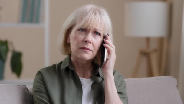Caucasian old woman mature middle-aged 50s offended lady aged businesswoman sitting on sofa at home listening to voice on phone talking smartphone receiving bad news sadness worry upset losing failure - Imágenes, Vídeo