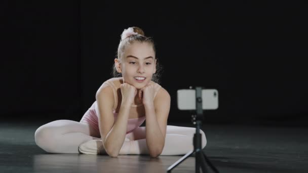 Young teen girl child teenager ballerina gymnast dancer sitting on floor recording vlog waving hello to mobile phone camera on tripod has online video call conference chat live broadcasting smartphone - Felvétel, videó