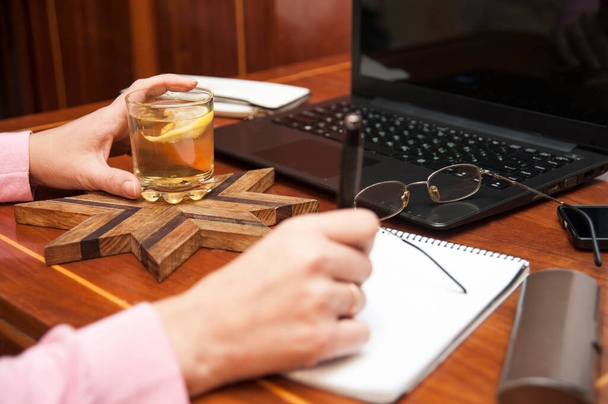 Een vrouw zit achter het bureau en werkt op een computer en schrijft iets in het notitieboekje en drinkt ook warme thee met citroen. Vrouw werkt thuis, bereidt een conferentie voor - Foto, afbeelding