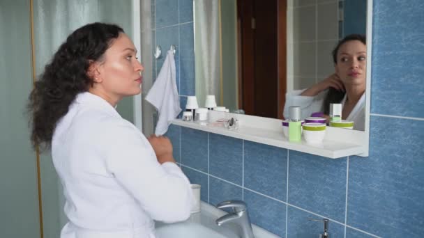 Dark haired beautiful multiethnic mature woman, wearing a white waffle bathrobe, combing hair, looking at her reflection in the bathroom mirror. Beauty and body care concept - Materiał filmowy, wideo