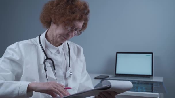 Senior woman doctor examines medical tests and patient exams in hospital office. Female doctor is holding a clipboard with medical documents and thinking about the diagnosis. Medicine and health care - Imágenes, Vídeo