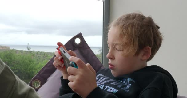 Dospívající chlapec tráví volný čas doma a hraje videohry na ruční konzoli. Moře a zelená příroda v okně - Záběry, video