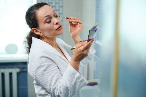 Beautiful middle-aged Hispanic woman in white bathrobe, holding a colorful eyeshadow palette and looking at her reflection in mirror while applying makeup on upper eyelids. Beauty and Fashion concept - Foto, afbeelding