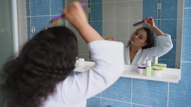 Pretty woman standing in front of mirror at bathroom and combing hair, caring for curly chevelure, during morning routines. Lifestyle, beauty care concept. Close-up shot. Hair Care Cosmetics Advert. - Materiał filmowy, wideo