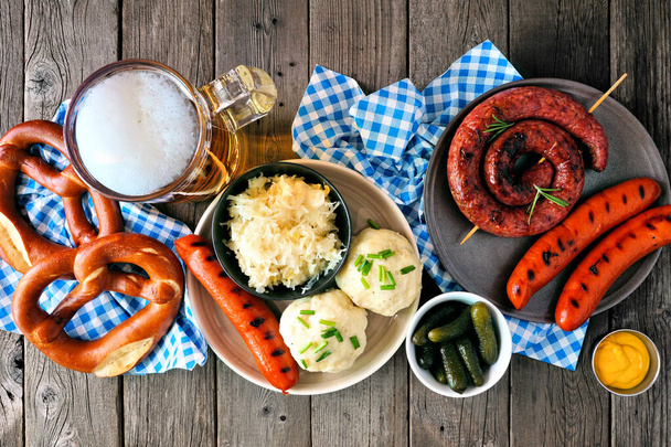Classic German Oktoberfest meal. Above view table scene over a dark wood background. Beer, sausage, pretzels, sauerkraut, and potato dumplings. - Фото, зображення