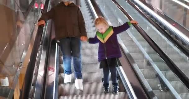 Brother and sister descend on the escalator. The brother is holding his sisters hand. The boy has a protective mask on his face. Children are dressed in warm jackets - Footage, Video