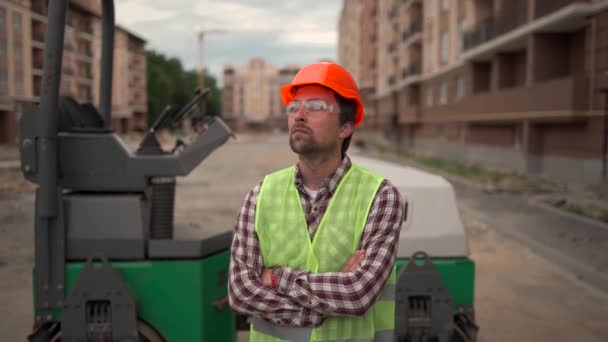Portrait of male construction worker with his hands on his chest proudly looking at result of construction and interjecting work done at construction site against the background of a paver machine.  - Video