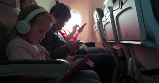 Mom, son and daughter are traveling by plane. They hold gadgets in their hands, a little blonde girl has white headphones on her head - Imágenes, Vídeo