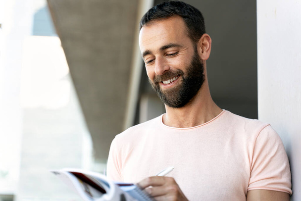 Portrait of smiling latin student studying, learning language, exam preparation, education concept. Handsome bearded hispanic man reading book, taking notes, standing on urban street   - Zdjęcie, obraz