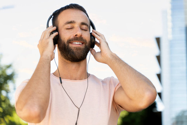 Smiling hispanic man listening music with eyes closed standing on the street. Portrait of handsome bearded hipster wearing headphones using modern technology outdoors  - Fotó, kép