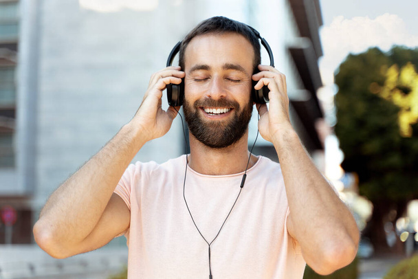 Usmívající se hispánec poslouchající hudbu se zavřenýma očima stojící na ulici  - Fotografie, Obrázek