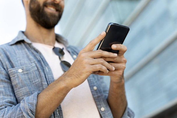 Lähikuva liikemiehen kädet käyttämällä mobiilisovellus ostoksia verkossa, lähettämällä tekstiviestin ulkona. Hymyilevä parrakas hipster tilalla älypuhelin pelaa mobiilipeli. Teknologian käsite   - Valokuva, kuva