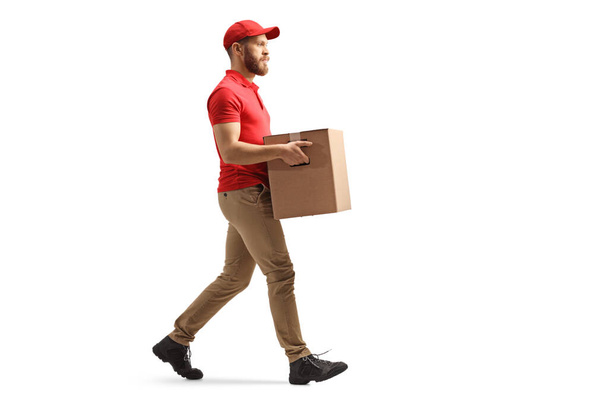 Full length profile shot of a courier carrying a cardboard box and walking isolated on white background - Foto, afbeelding