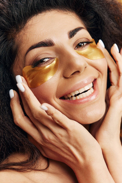 Iloinen ruskettunut melko latinalaisamerikkalainen nainen touch kulta hydrogeeli laastaria silmän alla nauttia ihonhoito menettely aiheuttaa eristetty keltaisella pohjalla. Kosmeettinen tuote mainos Natural kauneus käsite Studio muotokuva - Valokuva, kuva