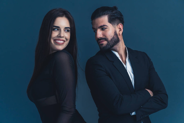 Young couple in black formal clothes standing back to back looking at each other flitring. Dark background studio shot. High quality photo - Photo, Image