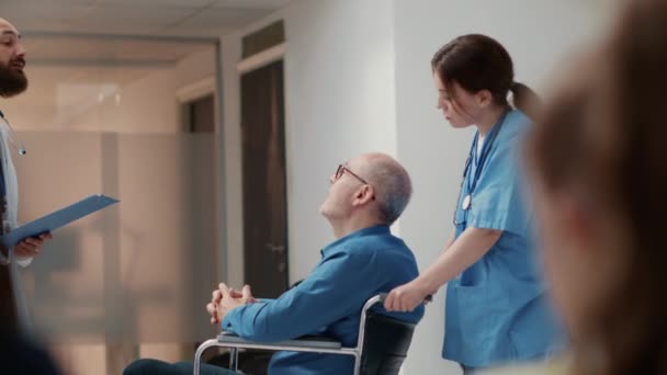 Nurse helping senior man with chronic disability to attend checkup visit with general practitioner. Retired wheelchair user with impairment in hospital reception lobby waiting room. Handheld shot. - Materiał filmowy, wideo