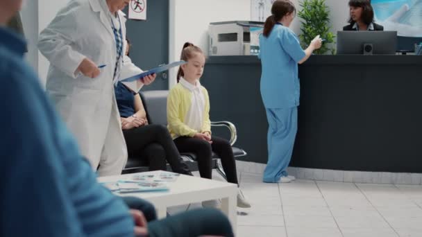 Porträt eines kleinen Mädchens und einer Mutter, die im Wartebereich einer medizinischen Klinik sitzen und auf einen Arzttermin beim Hausarzt warten. Krankenhausempfang mit vielfältigen Patienten. - Filmmaterial, Video