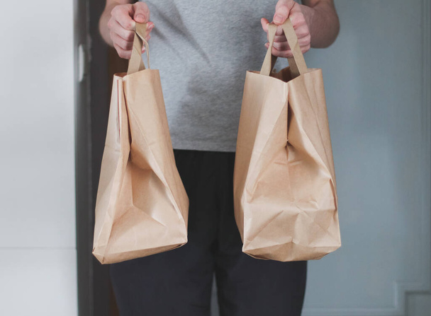 The hands of a caucasian young man hold out two paper craft bags with fastwood inside against the background of the entrance doors to the apartment, side view close-up with depth of field. Concept of food delivery, fast food, eco-friendly. - Foto, Imagen