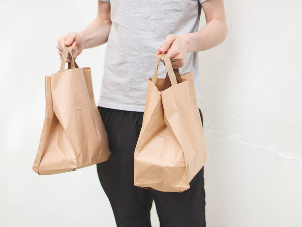 The hands of a caucasian young man in a gray t-shirt and black sweatpants hold out two paper craft bags with fastwood inside against the background of a white old wall in the backyard of the house, side view close-up with depth of field. Concept of f - Fotoğraf, Görsel