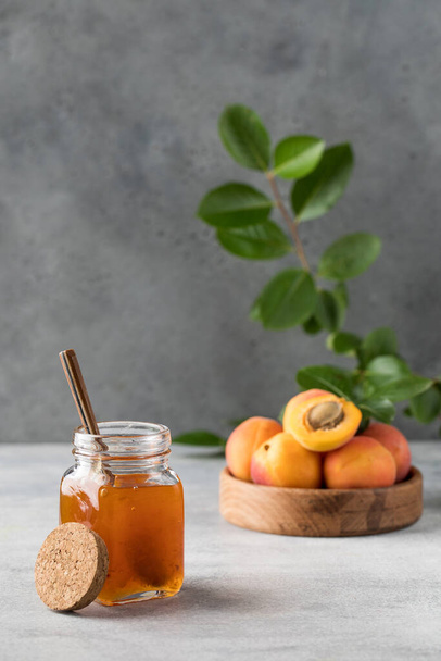 A jar with apricot jam and a spoon close-up on a gray background. In the background is a plate with fresh apricots and a branch with green leaves. Fruit preservation. - Photo, image