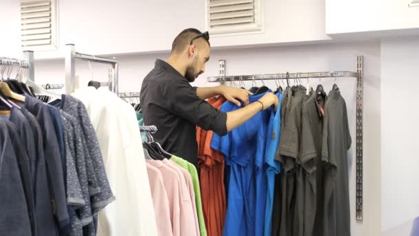 The young man who chooses what he likes from the colorful t-shirts in the aisle for buy, to examine the colorful clothes hanging on the hanger one by one - Materiał filmowy, wideo