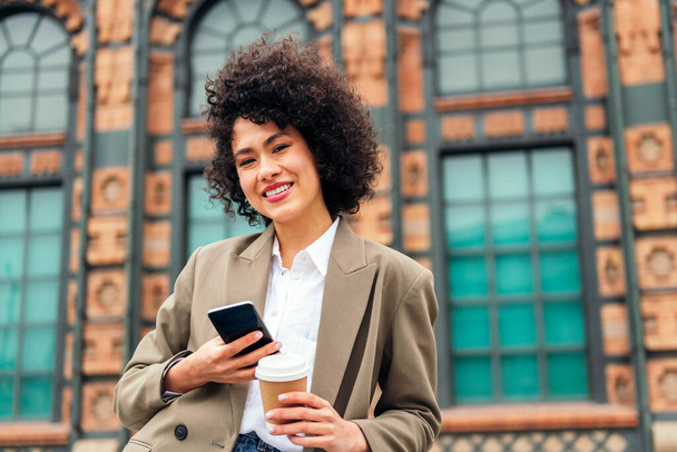 young business woman smiles happy looking at camera with a mobile phone in one hand and a coffee in the other, concept of urban lifestyle and technology, copy space for text - Foto, imagen