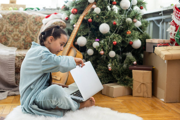 winter holidays, technology and children concept - little student girl with laptop computer at home over christmas tree background - Photo, Image