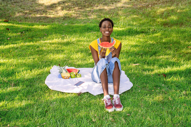 Very happy smiling black African woman sitting in an outdoor park while eating a watermelon and more fruit. High quality photo. - Фото, зображення