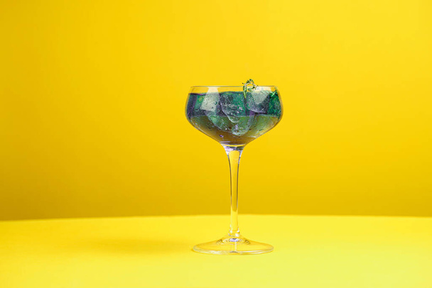 Dark blue drink in a vintage glass for sparkling wine - pea flower tea or blue curacao sirup cocktail with thyme branches on yellow background. Copy space for text - 写真・画像