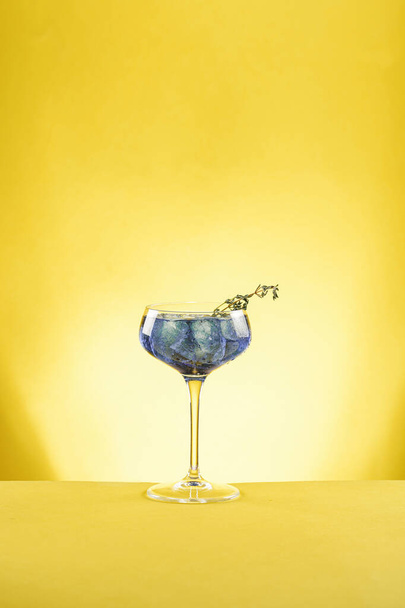 Dark blue drink in a vintage glass for sparkling wine - pea flower tea or blue curacao sirup cocktail with thyme branches on yellow background. Copy space for text - Foto, afbeelding