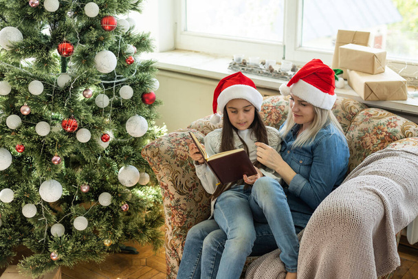 Pretty young mother reading a book to her daughter near Christmas tree. - Foto, Bild