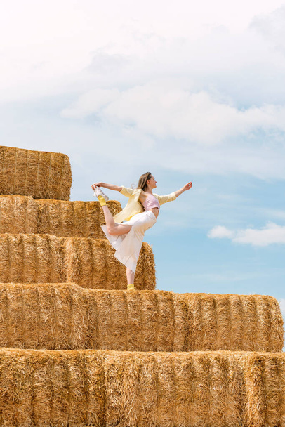 Young woman dances on huge block of hay. Hay harvesting for autumn. Girl practicing yoga on haystack - 写真・画像