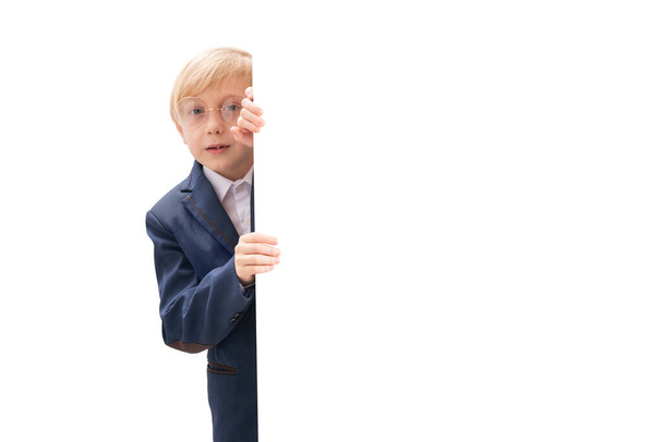 Portrait of blond little boy with white blank isolated on white background. Boy with glasses and school uniform peeking out from behind board - Foto, immagini