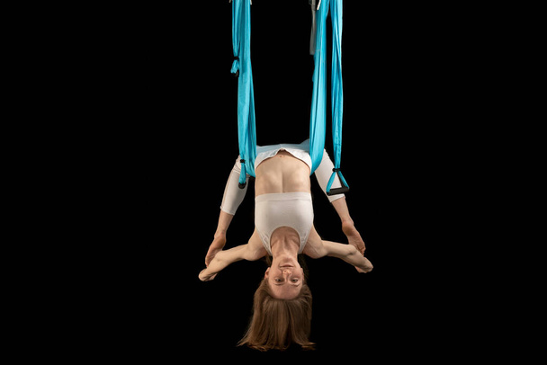 Girl in white clothes does gymnastic tricks on blue canvas. Young woman performing antigravity yoga exercise. - 写真・画像