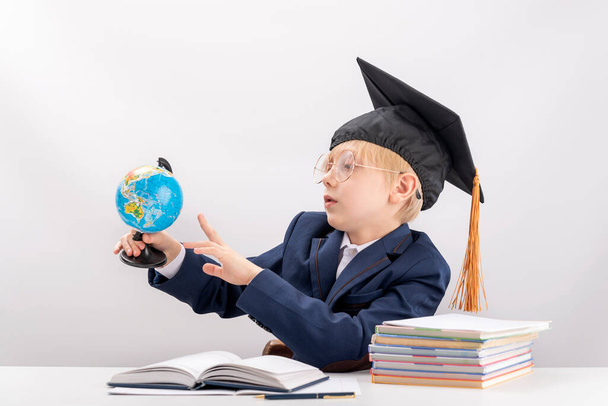 Okul üniformalı bir çocuk ve elinde dünya ile gezen bir öğrenci şapkası var. Kitapların ve defterlerin yanında. Okul çocuğu coğrafya okuyor.. - Fotoğraf, Görsel