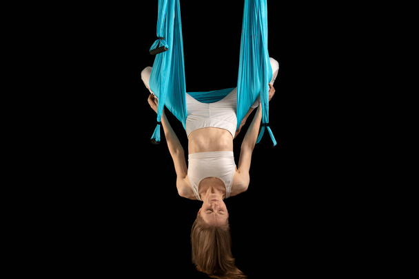 Young girl in white sportswear does fly yoga and stretches. Black background. Woman using canvas hammock for air anti-gravity yoga - Fotoğraf, Görsel