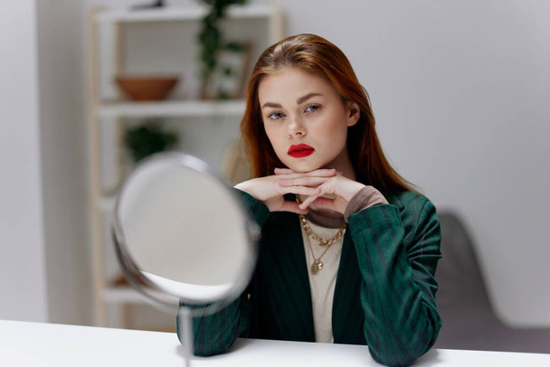 Mladá žena s rudými rty se připravuje na večerní kosmetické procedury doma před zrcadlem, které sedí u stolu v jasném pokoji. Koncept krásy v interiéru. Kvalitní fotografie - Fotografie, Obrázek