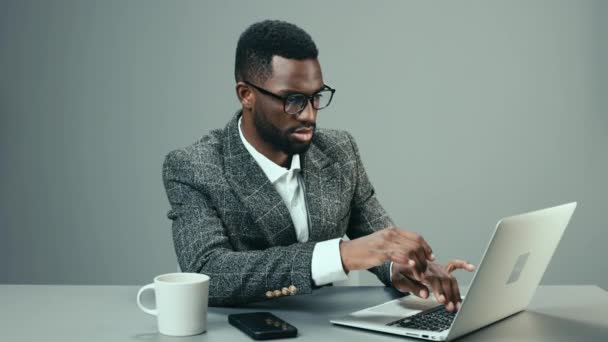 Uomo d'affari afroamericano in occhiali da computer lavora in ufficio dietro un computer portatile con una tazza di caffè alla sua scrivania su uno sfondo grigio ed è stanco. Filmati 4k di alta qualità - Filmati, video