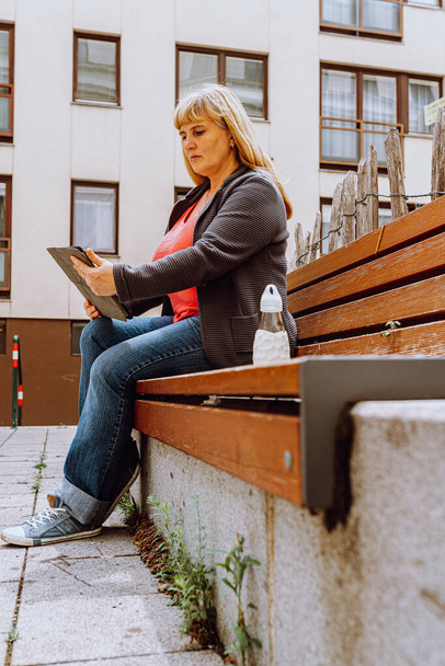 Mature happy middle-aged woman, 40s, with long blond hair, wearing jeans and knitted jacket, sitting on  bench in park, holding tablet in hands, video calling friend - Zdjęcie, obraz
