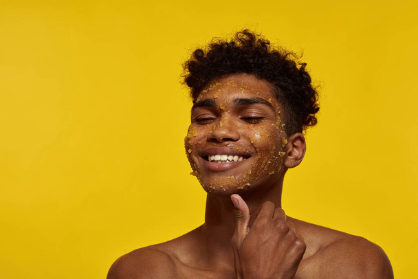 Partial of smiling black guy with honey scrub on his face skin. Young brunette curly man with closed eyes. Facial skin care. Isolated on yellow background. Studio shoot. Copy space - Fotó, kép