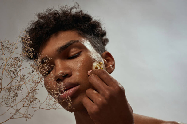 Cropped of black guy applying ice slice with plant inside on his face. Young man with closed eyes. Body and skin care. Concept of natural cosmetic. White brown background. Studio shoot. Copy space - 写真・画像